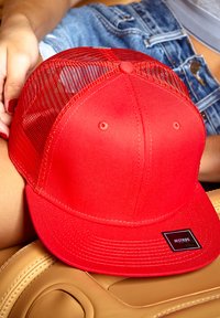 Rød baseballkasket med mesh-back, fladt skygge, broderet detaljer og et sort rektangulært mærke foran. Ligger på en tan læderoverflade.