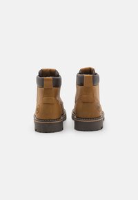 Botas de cuero marrón hasta el tobillo con un collar de tejido gris, tirador trasero y suela de goma texturizada. Detalles de costura a lo largo de la suela.