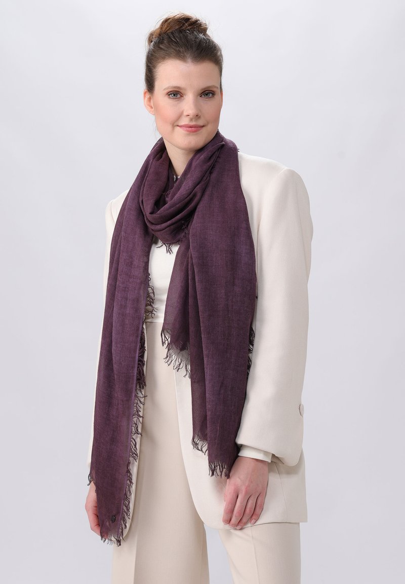 Sciarpa viola realizzata in un tessuto leggero, con bordo frangiato. Indossata avvolta attorno al collo, complementando un blazer color crema.