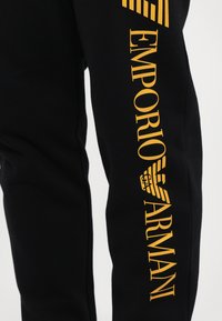 Čierne nohavice z mäkkej tkaniny s veľkým žltým logom "EMPORIO ARMANI" pozdĺž pravej nohy, s textúrovanými detailmi a moderným strihom.