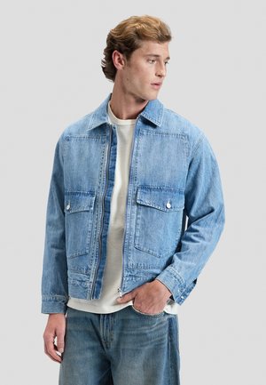 Ung mand iført lysblå denimjakke og jeans, der kigger til venstre med den ene hånd i lommen mod en ensfarvet grå baggrund.