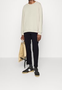 Man som bär en beige långärmad skjorta, svarta byxor, svarta sneakers med vita fläckar, och håller en beige tote-väska med svarta remmar.