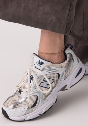 Weiße und silberne New Balance Laufschuhe, getragen mit brauner Hose und einem dünnen goldenen Fußkettchen.