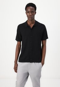 Black short-sleeve button-up shirt with a collared neckline and textured fabric. Paired with light gray pants. Simple design, casual fit.