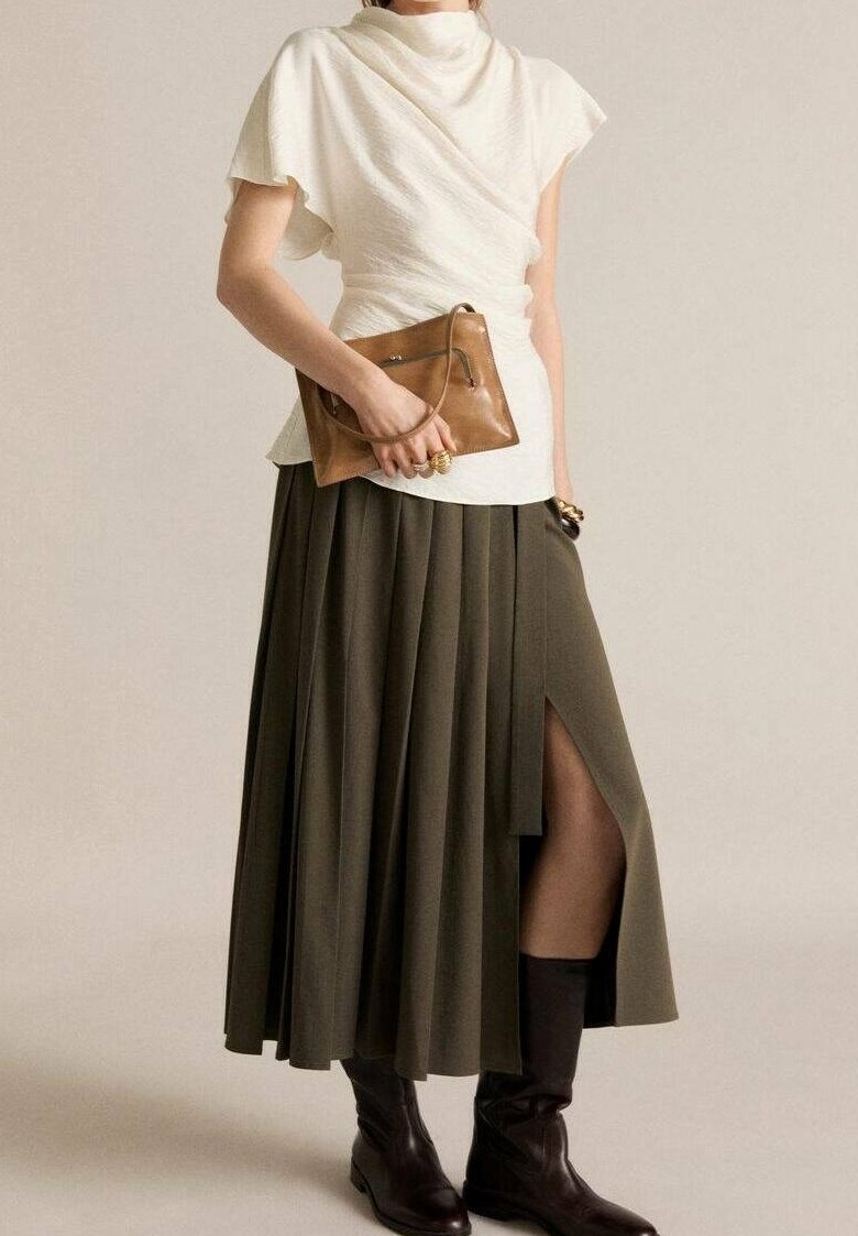 Femme en top blanc drapé, jupe plissée vert olive avec une fente latérale, bottes marron foncé, tenant une pochette en cuir marron.