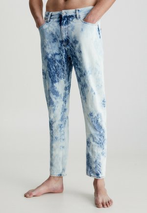 Person trägt hellblaue und weiße Acid-Wash-Jeans, steht barfuß mit den Händen in den Taschen vor einem schlichten Hintergrund.