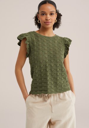 Frau trägt ein olivgrünes, gehäkeltes ärmelloses Oberteil mit Rüschen an den Schultern und beige Hosen mit Kordelzug, steht mit den Händen in den Taschen.