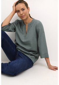 Top a maniche corte, verde oliva, con scollo a V e vestibilità ampia, abbinato a jeans in denim blu scuro. Tessuto semplice e strutturato.