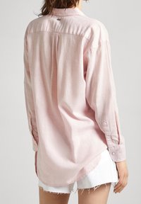 Langarm, lichtroze shirt gemaakt van zachte stof. Bevat een kraag, manchetten en een rugplooitje, met een gebogen zoom en minimale details.