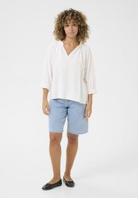 Blusa bianca leggera con scollatura a V e dettagli arricciati, abbinata a pantaloni shorts in denim e ballerine nere, che mettono in risalto un design semplice e uno stile casual.