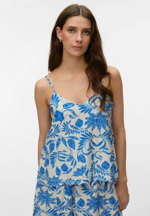 Vrouw draagt een beige hemdje en korte broek met blauwe bloemenprint, lang golvend bruin haar, staande tegen een effen witte achtergrond.