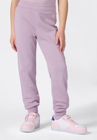 Person in hellblauen Jogginghosen und weißen sowie rosa Sneakers, vor einem schlichten weißen Hintergrund stehend.