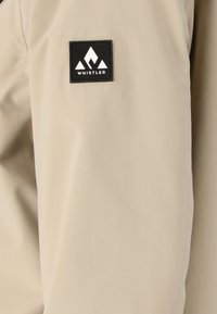 Beige vattentät jacka med en slät textur, med en svart fyrkantig logotyp med en bergsdesign på överarmen.