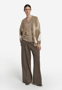 Beżowy sweter na guziki z dzianiny z wzorem diamentu, pod nim brązowy sweter z dzianiny oraz szerokie brązowe spodnie o fakturowanym wykończeniu.