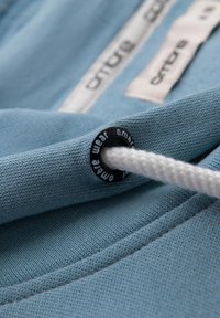 Tessuto azzurro chiaro con una superficie texturizzata, dotato di un occhiello in plastica nera e di un laccio bianco, include un'etichetta del marchio sullo sfondo.