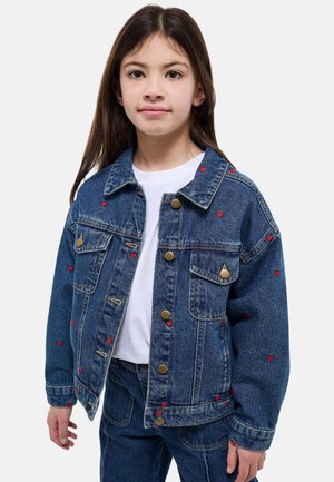 Fille portant une veste et un pantalon en denim bleu avec de petits motifs de cœurs rouges, et un t-shirt blanc uni, debout devant un fond blanc.