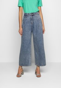 Vaaleansiniset, leveälahkeiset farkut, joissa on etupleissut ja keskipitkä vyötärö. Yhdistetty vihreään T-paitaan ja musta-valkoisiin raidallisiin korkokenkiin.