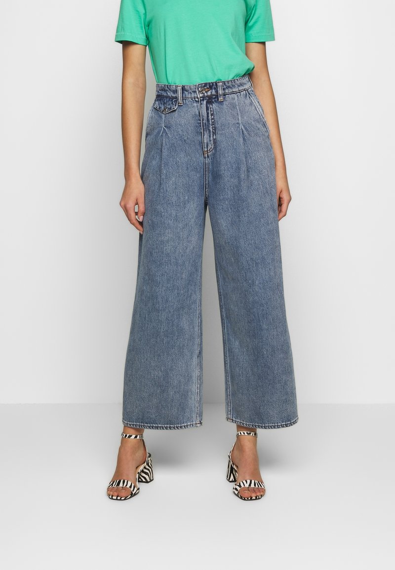 Vaaleansiniset, leveälahkeiset farkut, joissa on etupleissut ja keskipitkä vyötärö. Yhdistetty vihreään T-paitaan ja musta-valkoisiin raidallisiin korkokenkiin.