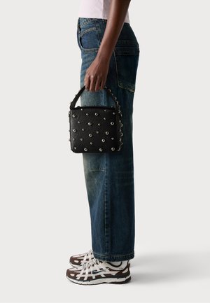 Persona che indossa jeans blu e sneakers bianche, mentre tiene una borsa nera con borchie per il manico superiore contro uno sfondo semplice.