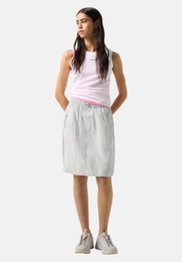 Mujer con cabello largo y oscuro que lleva una camiseta sin mangas rosa y blanca, falda gris clara con bolsillos con cremallera y zapatillas blancas, de pie con las manos detrás.