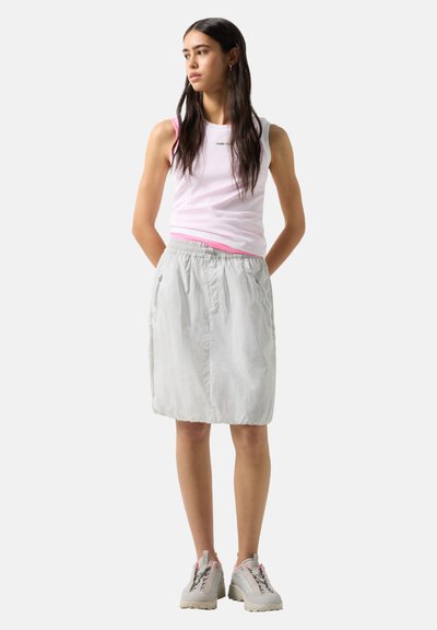 Femme aux longs cheveux foncés portant un top sans manches rose et blanc, une jupe gris clair avec des poches zippées, et des baskets blanches, debout les mains derrière.