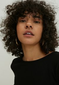 Schwarzes geripptes Oberteil mit einem Bootsausschnitt, das eine Nahaufnahme des Gesichts des Models zeigt, mit lockigem Haar und goldenen Hängeohrringen. Neutrale Hintergrund.