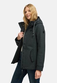 Nainen, jolla on pitkä vaalea hius, pukeutuneena tummanharmaaseen hupparintakkia, joka on avattu kevyen yläosan ja sinisten farkkujen päällä, hymyilee hieman vasemmalle.