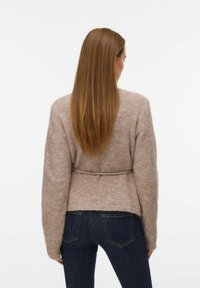 Svijetlosmeđi džemper sa teksturom i pojasom u struku, dugim rukavima i opuštenim krojem, u paru s tamnoplavim trapericama.