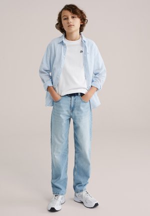 Chemise à rayures bleues et blanches boutonnée sur un T-shirt blanc, associée à un jean bleu clair et des baskets blanches. Mains dans les poches, posture debout.