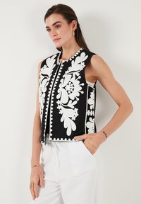 Zwarte mouwloze vest met witte bloemmotieven en pompoms, met een rechte snit en een sluiting aan de voorkant. Lichte stoffen textuur.