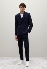 Blazer navy doppiopetto con quattro bottoni, pantaloni coordinati e sneakers bianche. Tessuto morbido, vestibilità su misura, design minimalista.