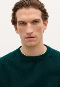 Donkergroen geribbelde trui met een ronde halslijn, textuurstof en subtiele details op de schouders, gedragen over een licht shirt.