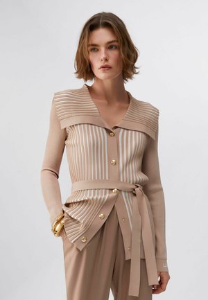 Donna che indossa un cardigan a righe beige e bianche con bottoni, ampio colletto e cintura annodata, abbinato a pantaloni beige e braccialetti dorati.