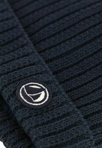 Maglione lavorato a maglia blu navy con texture a coste, dotato di un'etichetta ricamata rotonda in bianco e blu navy. Il tessuto sembra spesso e strutturato.