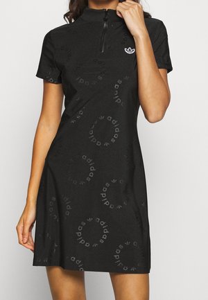 Kobieta w czarnej sukience Adidas z krótkim rękawem, z suwakiem przy kołnierzu i subtelnymi okrągłymi wzorami z logo Adidas.