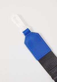 Etichetta in pelle sintetica blu con cuciture bianche attaccata a una cinghia di tessuto nera, dotata di un clip in plastica trasparente per il fissaggio.