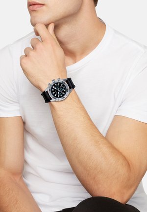 Schwarze Uhr mit silberner Hardware, ausgestattet mit einem runden Zifferblatt und mehreren Anzeigen. Das Armband besteht aus schwarzem Gummi, was den Komfort und die Haltbarkeit erhöht.