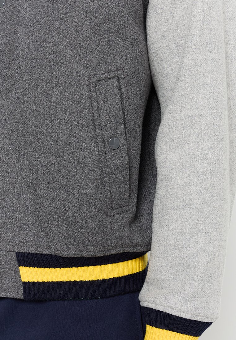 Veste grise et gris clair avec une poche boutonnée, un ourlet côtelé bleu marine et jaune, fabriquée dans un tissu doux et texturé.