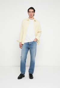 Μακρυμάνικο πουκάμισο με ανοιχτό κίτρινο ριγέ μοτίβο πάνω από λευκό T-shirt, συνδυασμένο με φθαρμένο μπλε τζιν και μαύρα παπούτσια. Απλός, casual σχεδιασμός.