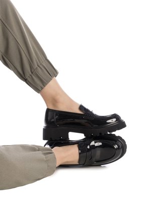 Mocasines de charol negro con suela gruesa, que presentan una correa decorativa y detalles de costura sutiles. Combinados con pantalones de color verde oliva.
