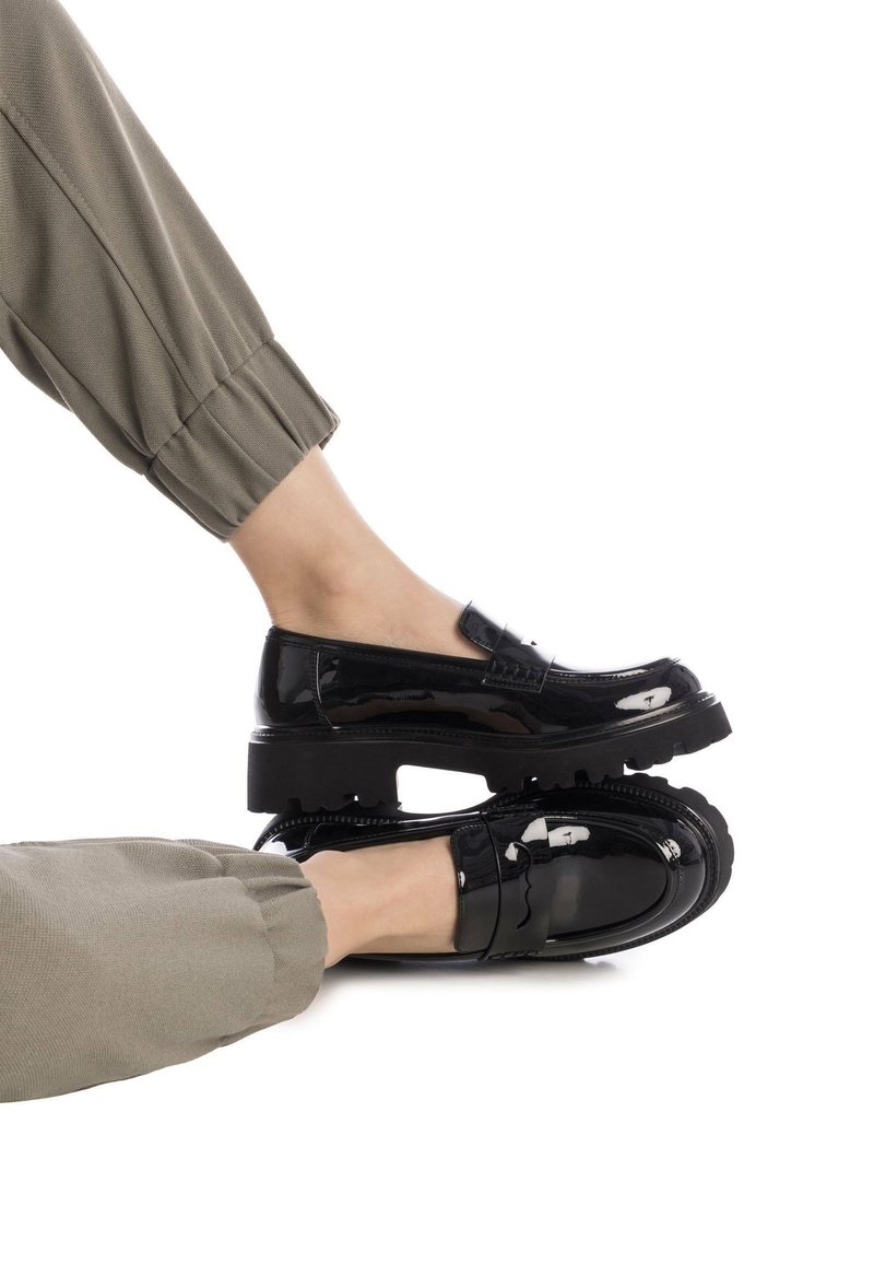 Mocasines de charol negro con suela gruesa, que presentan una correa decorativa y detalles de costura sutiles. Combinados con pantalones de color verde oliva.