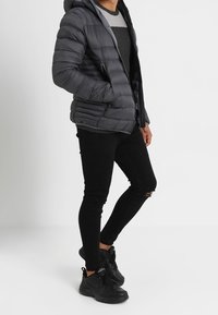 Doudoune noire à capuche avec un design matelassé, associée à un jean noir déchiré et des chaussures de sport noires.