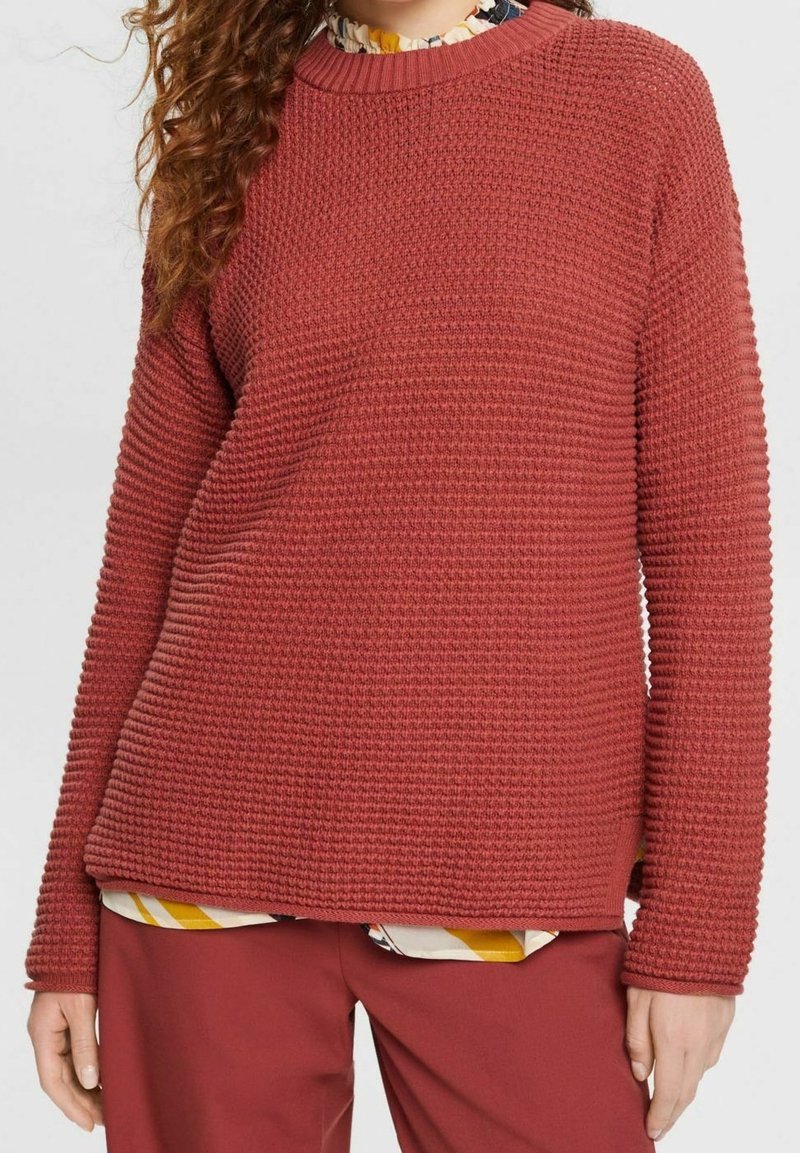 Femme portant un pull à manches longues rouge rouille texturé par-dessus un chemisier à motifs, assorti à un pantalon rouge rouille, visible des épaules jusqu'aux hanches.