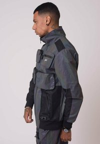 Giacca grigia iridescente con accenti neri, dotata di molteplici tasche, colletto alto e chiusura con zip. Presenta un tessuto testurizzato e un design aderente.