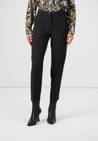 Femme portant un pantalon noir ajusté, des bottines à talons hauts noires et une chemise à manches longues à fleurs, se tenant devant un fond uni.