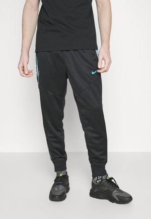 Personne portant un jogging Nike noir avec un logo bleu et des rayures, des baskets noires, une chemise noire, debout sur un sol et un fond gris clair.