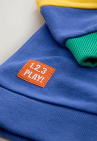 Bunt gestickter Pullover mit blauem Stoff, gelben Schultern und grünen Bündchen. Beinhaltet ein rechteckiges oranges Etikett mit "1.2.3 PLAY!" in weiß.