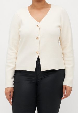 Kobieta w kremowym swetrze z dekoltem w serek na trzy guziki i czarnych spodniach, stojąca na tle jednolitego, białego tła.