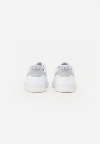 Hvide sneakers med grå detaljer, glat læderoverdel, flad hvid gummisål og "Stan Smith" logo på bagsiden. Minimalistisk design.