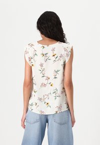 Nainen, jolla on pitkät kiharat hiukset, pukeutuneena valkoiseen ilmanhihoihin kukkablouseen ja vaaleansinisiin leveälahkeisiin farkkuihin, näkyy selkäpuolelta yksinkertaista taustaa vasten.